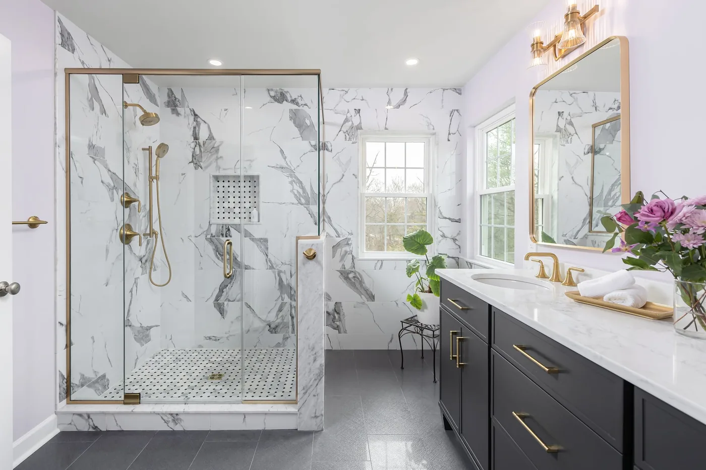 The same primary bathroom after the remodel, with a large frameless marble walk-in shower behind a half-wall, brushed brass fixtures, and a dual charcoal vanity with quartz tops.